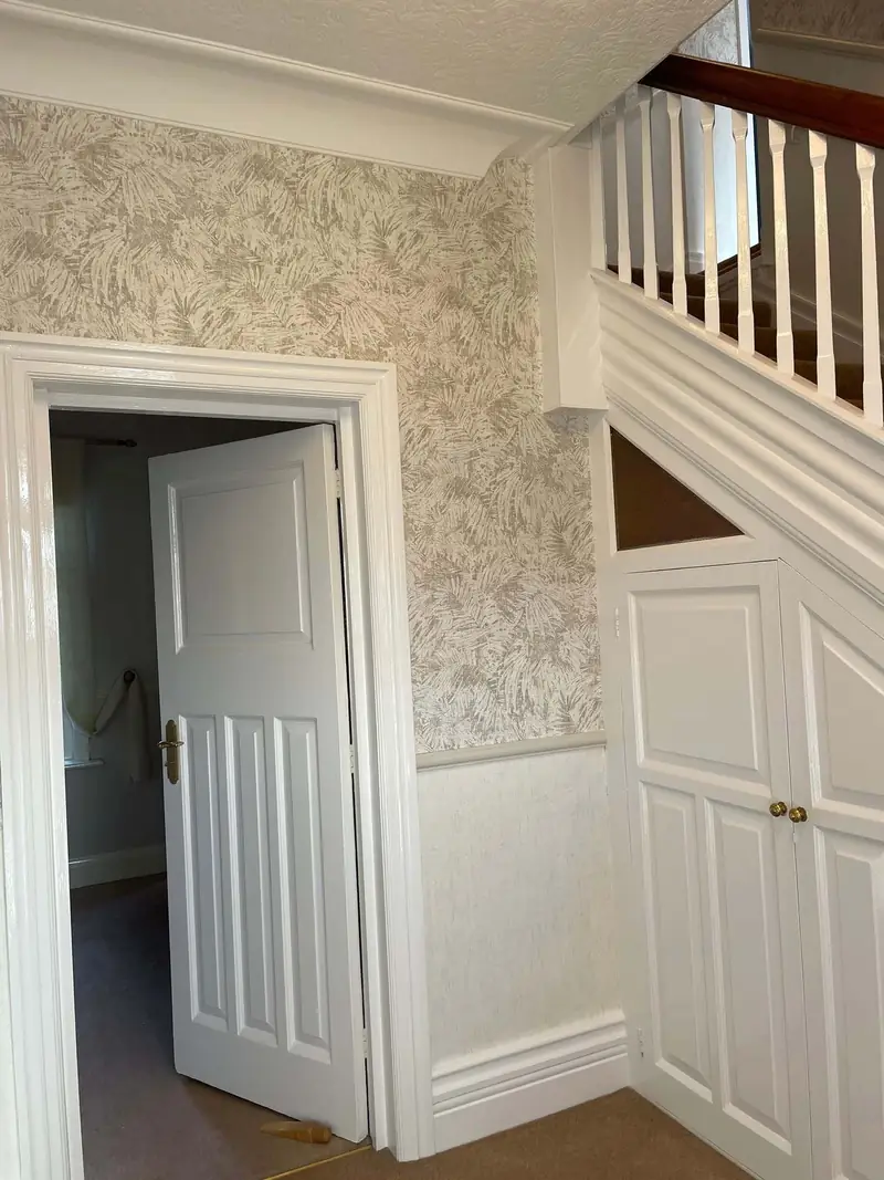 Hallway with fern-print wallpaper above a white dado rail, a white panelled door and a white understairs cupboard