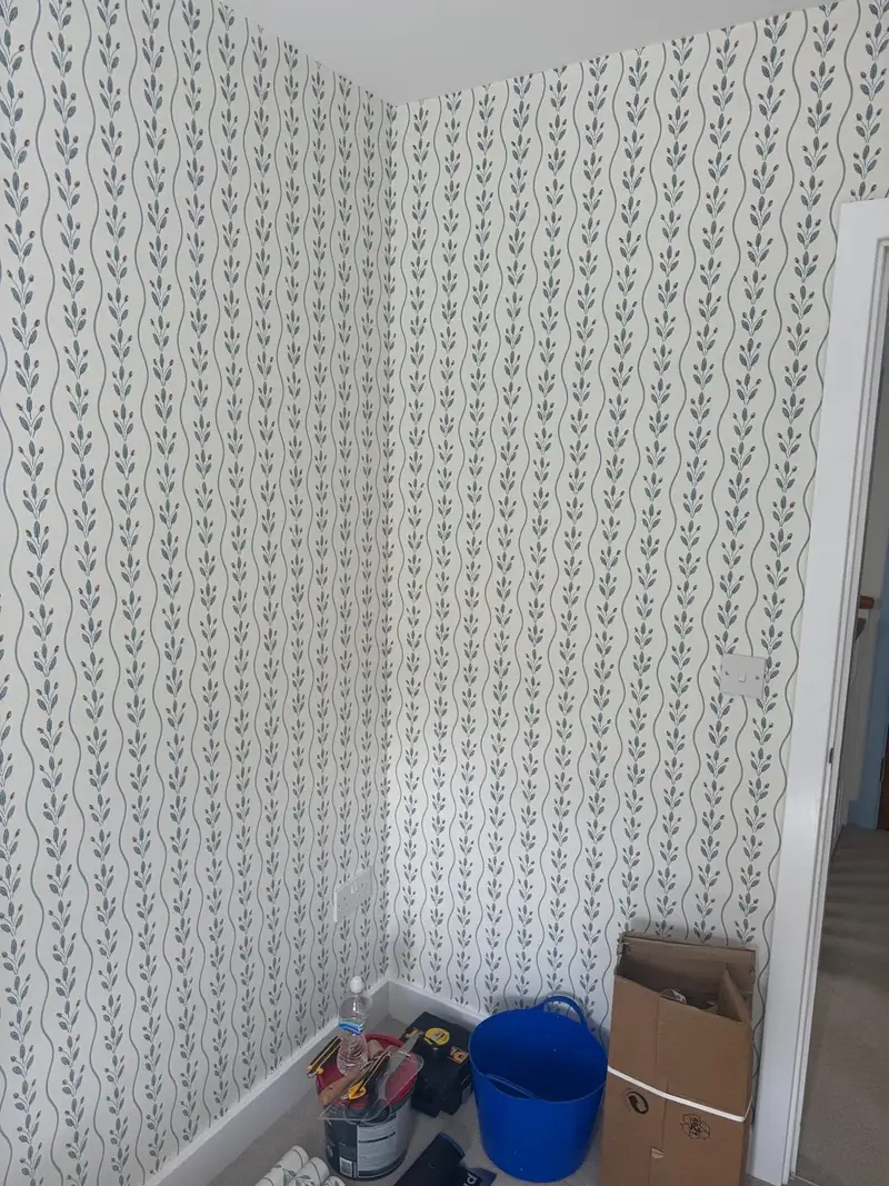 Small room papered on every wall in cream wallpaper with trailing blue leaves and red buds, decorating tools on the floor
