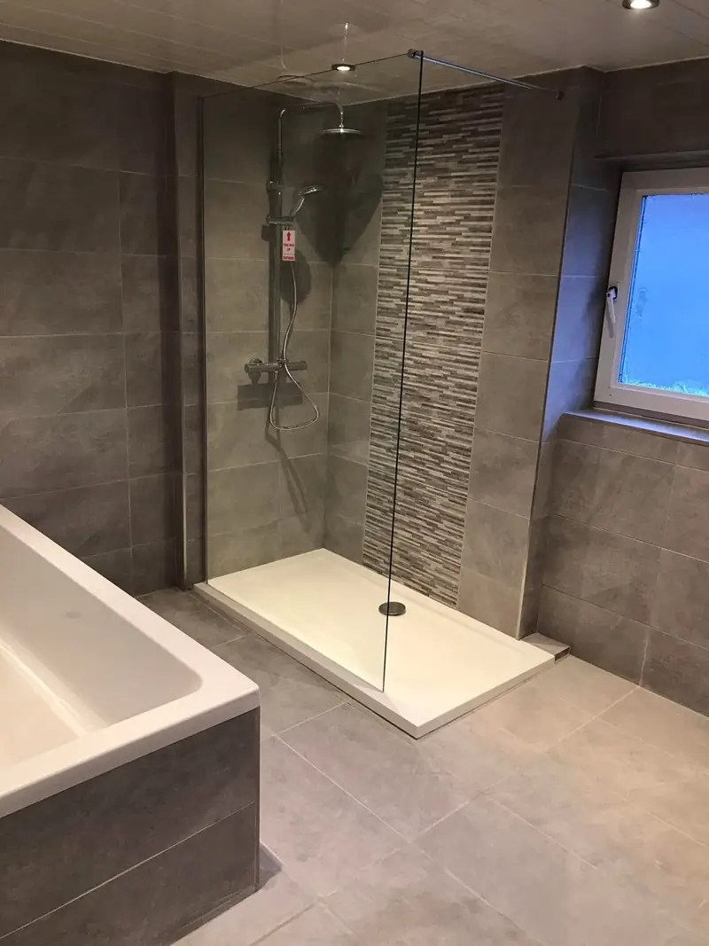 Walk-in shower with a glass screen and rainfall shower, grey wall tiles and a mosaic feature strip, beside a tiled bath